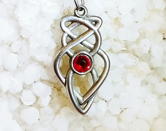 May include: A silver-toned pendant necklace with a Celtic knot design and a round, red gemstone at the center. The pendant hangs from a delicate chain. The necklace is displayed on a white, textured surface against a deep red background.