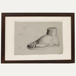 May include: A framed charcoal drawing on light grey paper depicting a side view of a human foot resting on a flat base. The artwork features detailed shading and anatomical contours, enclosed within a dark brown wooden frame with a white mat border.