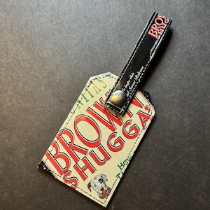 May include: A luggage tag with the words "BROWN SHUGGA" in red letters on a cream background. A dog illustration is in the lower right corner. The tag has a black strap with the word "BROWN SHUG" in red.