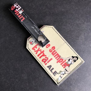 May include: A beige luggage tag with black stitching and a black leather strap. The tag features a cartoon illustration of a woman in a red and white outfit with the text "Sumpin Extra! ALE" in red and black.