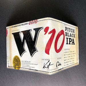 Puede incluir: Una cartera de cuero blanco con un diseño negro y rojo. La cartera presenta el texto "Brewmasters Release: 2010", "W'10", "Pitch Black IPA", y "Gold Medalist".