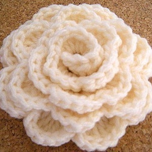 May include: A crocheted flower in a cream color. The flower is made of multiple layers of crocheted fabric, creating a three-dimensional effect.