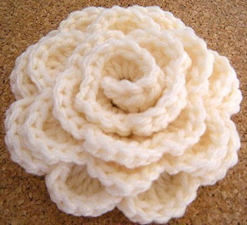 May include: A crocheted flower in a cream color. The flower is made of multiple layers of crocheted fabric, creating a three-dimensional effect.