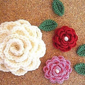 May include: Three crocheted flowers with pearl centers. The flowers are white, pink, and red. The flowers are surrounded by green crocheted leaves.