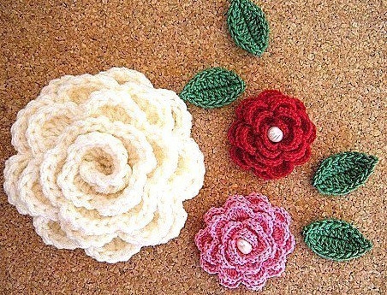 May include: Three crocheted flowers with pearl centers. The flowers are white, pink, and red. The flowers are surrounded by green crocheted leaves.