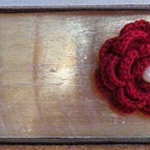 May include: A red crocheted flower with a white pearl button in the center. The flower is attached to a book.
