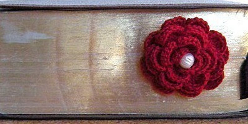 May include: A red crocheted flower with a white pearl button in the center. The flower is attached to a book.