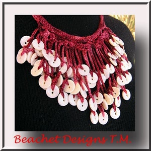 May include: A burgundy crocheted necklace with a fringe of white buttons. The necklace is displayed on a black background.