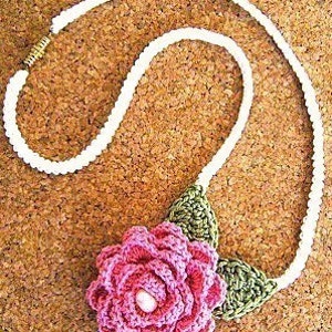 May include: A white crocheted necklace with a pink crocheted flower and green crocheted leaves. The flower has a small pink bead in the center.
