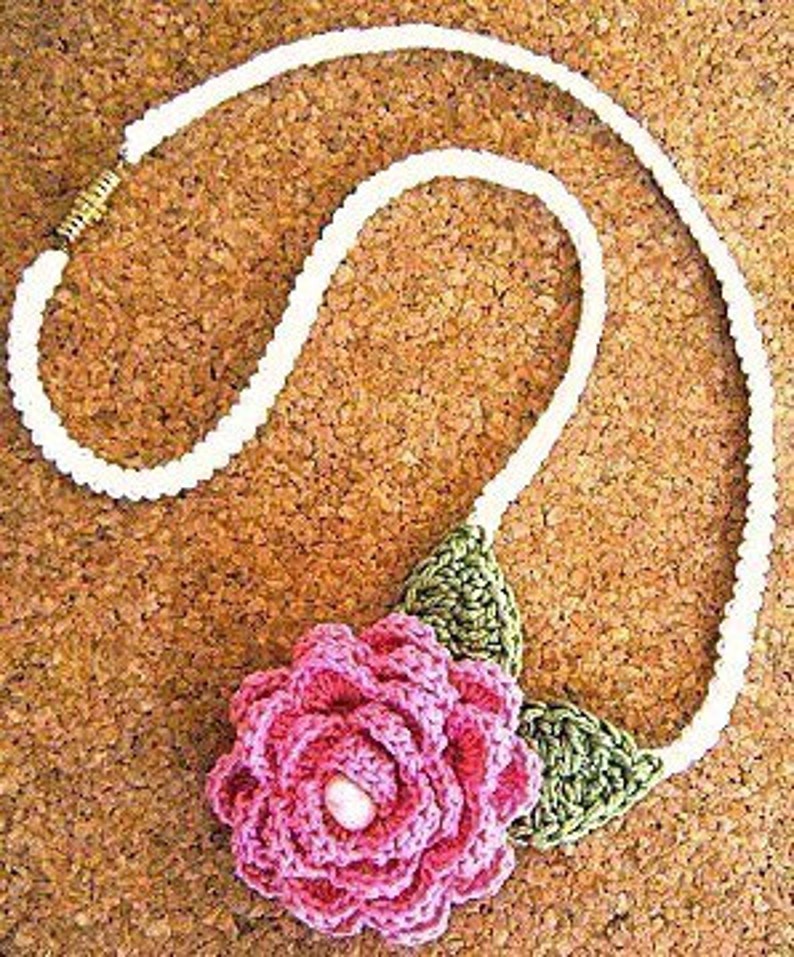 May include: A white crocheted necklace with a pink crocheted flower and green crocheted leaves. The flower has a small pink bead in the center.