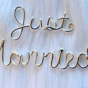 May include: Silver wire sculpture that reads "Just Married" on a white fuzzy background.