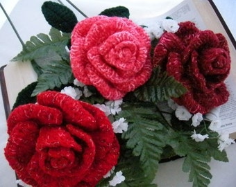May include: Three crocheted roses in shades of red and pink, arranged on a book with white flowers and green leaves.