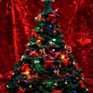 May include: A handcrafted Christmas tree made of green yarn, adorned with red bows, ornaments, and twinkling lights. A star tops the tree, set against a red velvet backdrop.