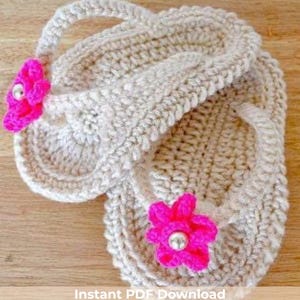 Könnte beinhalten: Ein Paar handgefertigte, beige gehäkelte Flip-Flops. Jeder Flip-Flop hat eine leuchtend rosa Blume mit einem silbernen Perlenmittelpunkt. Die Flip-Flops haben ein strukturiertes Aussehen und werden auf einer Holzoberfläche präsentiert.