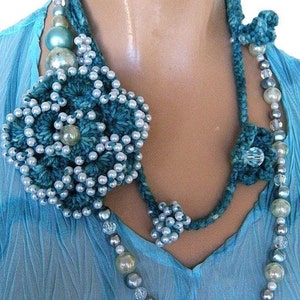 Pode incluir: Um colar de crochê azul turquesa com um grande pingente de flor. O colar é feito com contas e cristais em tons de turquesa, azul e prata.