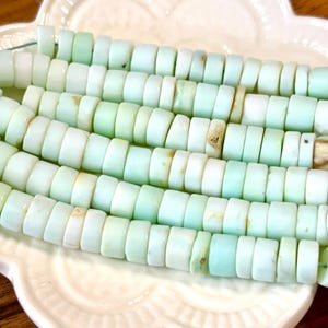 May include: A strand of light green and white opal beads. The beads are round and have a smooth, polished finish. They are strung on a clear thread and are arranged in a row.