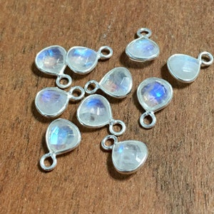 May include: A collection of nine teardrop-shaped moonstone charms with a silver-colored bezel and a small loop at the top. The moonstones display a shimmering, iridescent play of color, with hues of blue, purple, and white. The charms are set against a warm, brown background.