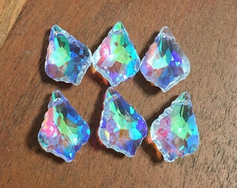 May include: Six iridescent crystal chandelier pendants with a faceted, teardrop shape. The crystals reflect a rainbow of colors, including pink, green, and blue. The pendants have a rough, textured edge and are arranged on a wooden surface.