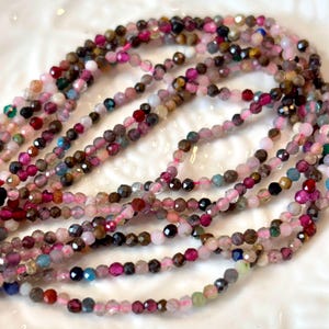 May include: Strands of faceted gemstone beads in a variety of colors, including pink, red, brown, and blue. The beads are strung on a pink cord, creating a delicate and colorful jewelry component.