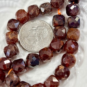 May include: Close-up of a strand of faceted, reddish-brown garnet beads. The beads are square-shaped and strung on an orange thread. A US quarter coin is included for size reference. The beads have a sparkling, polished appearance.