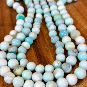 May include: Strands of round, polished beads in shades of pale blue, turquoise, and white. The beads have a smooth, glossy finish and vary slightly in color and pattern. The beads are strung together, creating a necklace or bracelet.