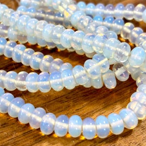 May include: Strands of opalescent beads in varying shades of white and blue. The beads are round with a slightly flattened shape, strung together to form multiple rows. The beads have a translucent quality, with a subtle shimmer. The background is a warm, wooden surface.