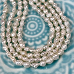 May include: A strand of white freshwater pearls, with an irregular, slightly elongated shape. The pearls are strung together on a clear thread and are displayed on a blue and white patterned surface.