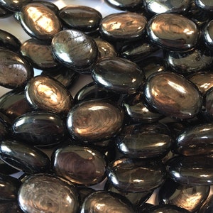 May include: A close-up of a pile of black obsidian gemstone beads. The beads are oval shaped and have a shiny, iridescent finish.