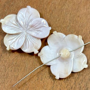 May include: Two white mother of pearl flower shaped beads with a silver wire threaded through the holes. The beads are carved with a detailed flower design.