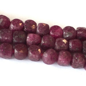 May include: A strand of deep red ruby beads, each bead is faceted and has a slightly irregular shape. The beads are strung on a thin thread.