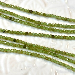 May include: Close-up of several strands of small, faceted, light green beads. The beads are strung together, creating a delicate and shimmering effect. The beads are a pale, spring green color.