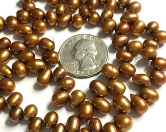May include: A collection of small, oval-shaped bronze-colored pearls arranged in a loose circular pattern around a silver-colored United States quarter coin. The pearls have a smooth, reflective surface and a metallic luster, contrasting with the coin.