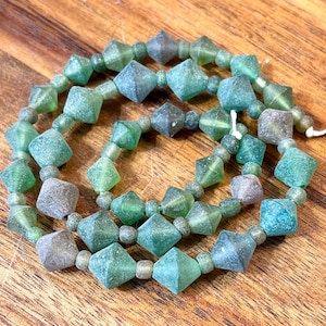 May include: Strands of green and gray glass beads. The beads are a mix of diamond and round shapes, strung together. The beads have a frosted appearance and are displayed on a wooden surface.