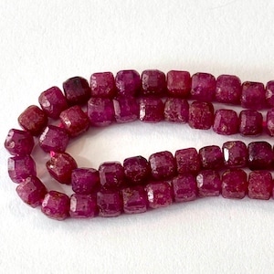 May include: Strands of faceted, deep red ruby beads. The beads are square-shaped with rounded corners and a slightly irregular surface. The beads are strung on a pink cord, and the background is white.