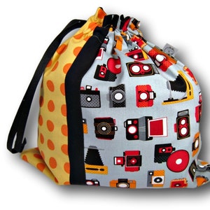 May include: A drawstring bag made of fabric with a blue background and a pattern of vintage cameras. The bag has a yellow and orange polka dot side panel with black trim.