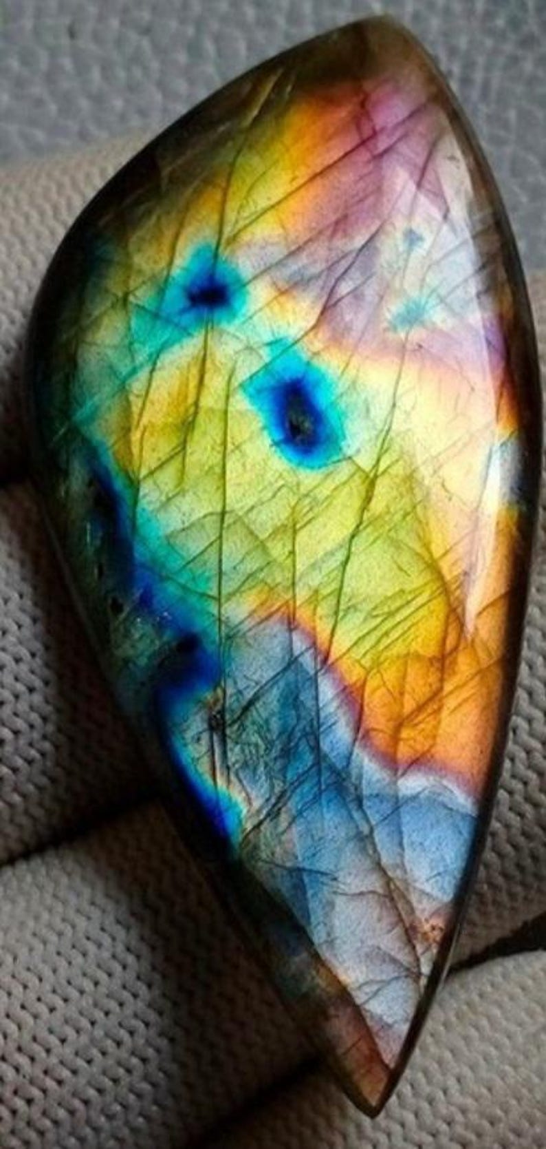 May include: A large, iridescent labradorite gemstone with a teardrop shape. The stone has a mix of blue, green, yellow, and orange colours.