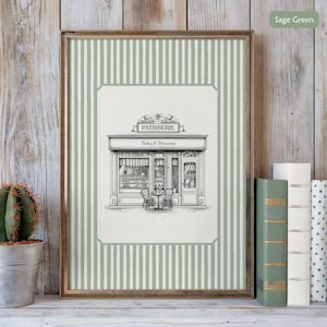 May include: A framed print featuring a black and white illustration of a patisserie shop with the word "PATISSERIE" above the entrance. The print has a sage green and white striped border and is displayed on a wooden shelf with books and a cactus.