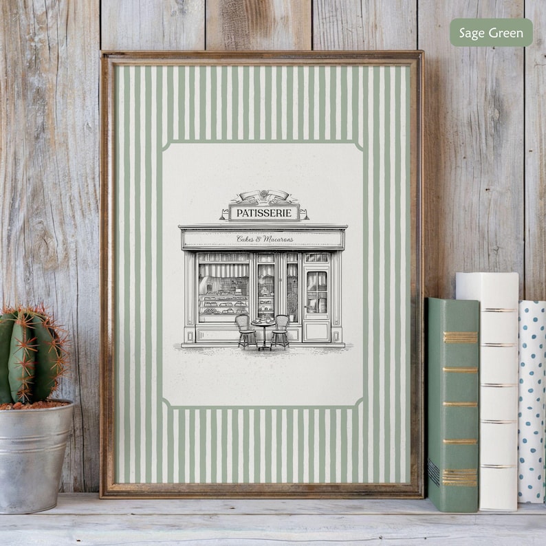 May include: A framed print featuring a black and white illustration of a patisserie shop with the word "PATISSERIE" above the entrance. The print has a sage green and white striped border and is displayed on a wooden shelf with books and a cactus.