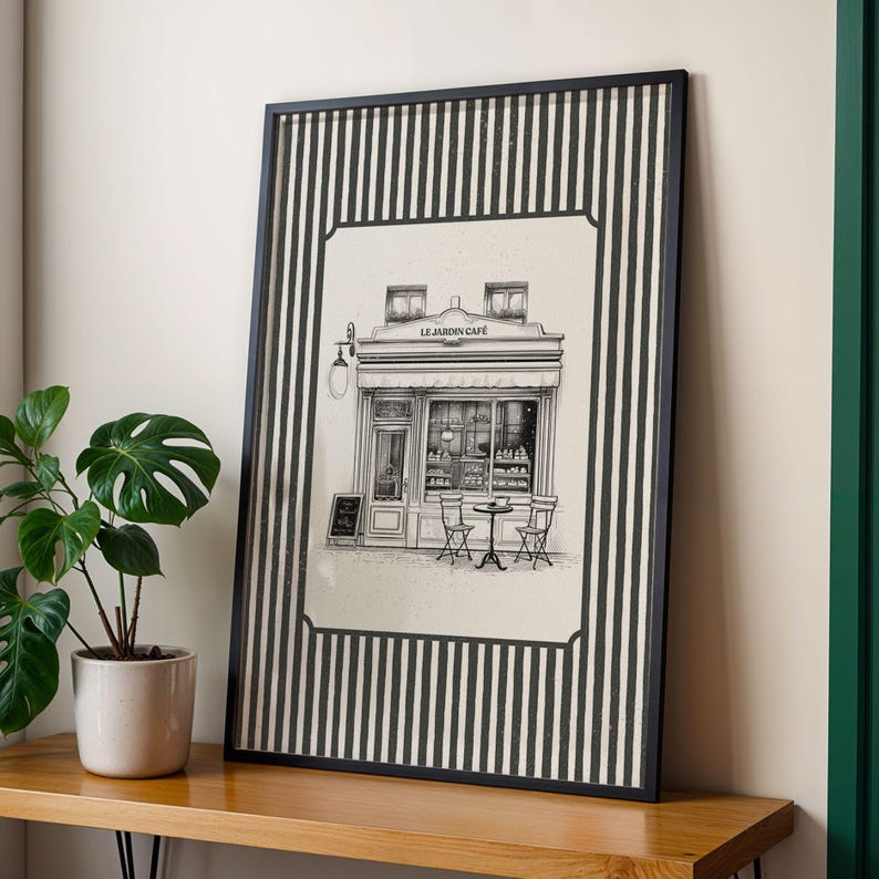 May include: Framed black and white print of a cafe, titled "Le Jardin Cafe". The artwork features a detailed illustration of a cafe exterior with tables and chairs. The frame has a black and white striped border.