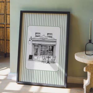 May include: Black framed print featuring a detailed black and white illustration of a cafe facade with the text "LE JARDIN CAF&Eacute;". The print has a green and white striped border and is leaning against a wall.