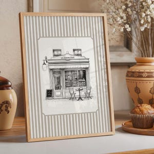 May include: A framed print featuring a black and white illustration of a cafe, "Le Jardin Cafe", with outdoor seating. The frame is light wood, and the background has vertical gray and white stripes. A muffin and vase are in the background.