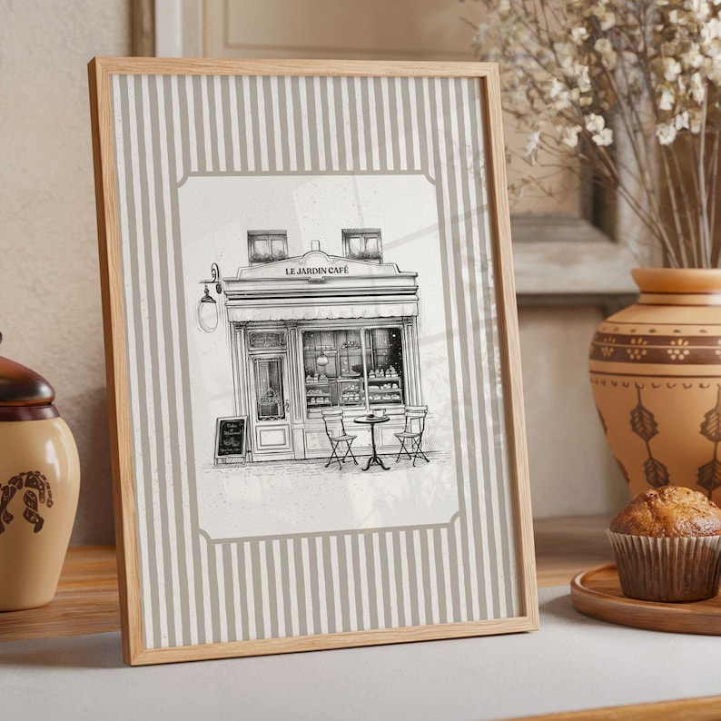 May include: A framed print featuring a black and white illustration of a cafe, "Le Jardin Cafe", with outdoor seating. The frame is light wood, and the background has vertical gray and white stripes. A muffin and vase are in the background.