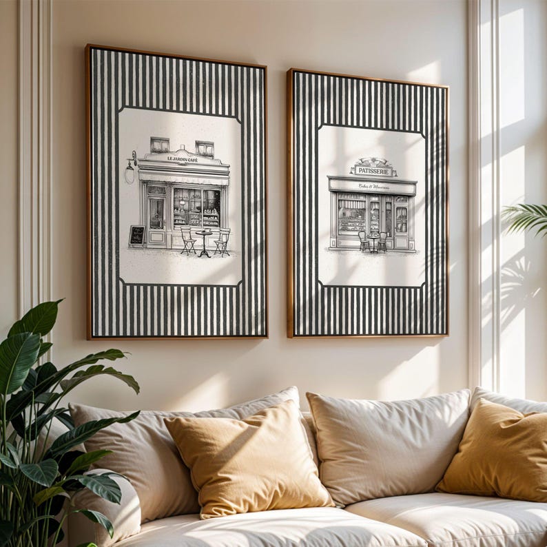 May include: Two framed prints of Parisian cafes. Each print features a black and white illustration of a cafe with outdoor seating, set against a background of black and white vertical stripes. The frames have a warm brown finish.