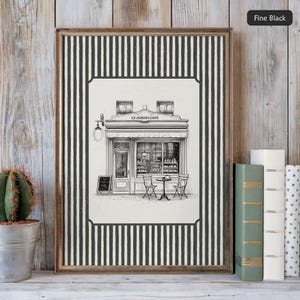 May include: Framed black and white print of a cafe, "Le Jardin Cafe", with a striped border. The print is displayed on a wooden shelf with books and a cactus in a metal pot. The cafe illustration shows tables, chairs, and a menu board.