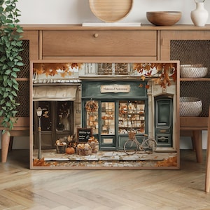 May include: A framed print depicts a quaint French bakery, "Maison d'Automne," with a teal facade, baskets of bread, and a bicycle. Autumn leaves and pumpkins add seasonal charm. The shop's window displays various baked goods.