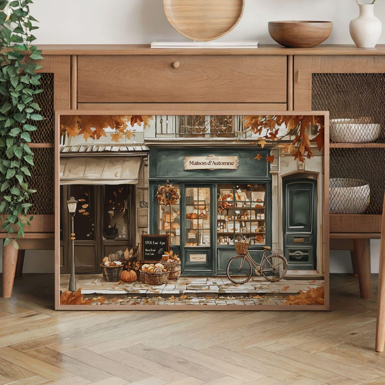May include: A framed print depicts a quaint French bakery, "Maison d'Automne," with a teal facade, baskets of bread, and a bicycle. Autumn leaves and pumpkins add seasonal charm. The shop's window displays various baked goods.