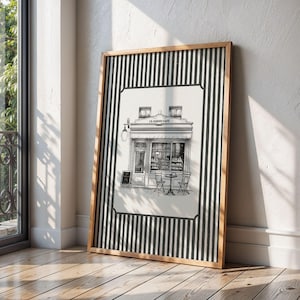 May include: A framed black and white print of a cafe, titled "Le Jardin Cafe". The print is in a wooden frame with a black and white striped border. The cafe has a detailed facade with tables and chairs outside.