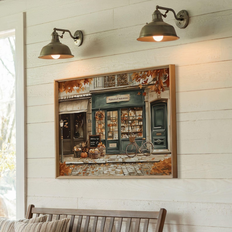 May include: A framed painting depicts a quaint shop, "Maison d'Automne," with a bicycle out front, pumpkins, and autumn leaves. Two antique-style wall sconces with dome shades are mounted above the painting on a white shiplap wall.
