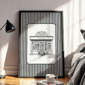 May include: Black and white framed print of a patisserie shop. The artwork features a detailed illustration of a bakery with a striped border. The shop's sign reads "Patisserie" above "Cakes & Macarons". A cup of coffee sits on a floral book.