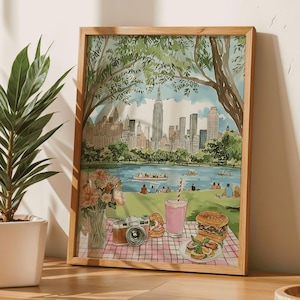 Könnte beinhalten: Ein gerahmtes Aquarellbild, das eine Picknickszene im Central Park in New York City zeigt. Das Bild zeigt eine Skyline der Stadt, einen See mit Booten, Menschen, die auf dem Gras picknicken, und einen Picknickkorb mit Essen und Getränken.